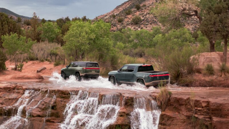 Two green Rivian Gen 2 off-road vehicles drive through shallow water at the edge of a small waterfall in a rocky, forested landscape.