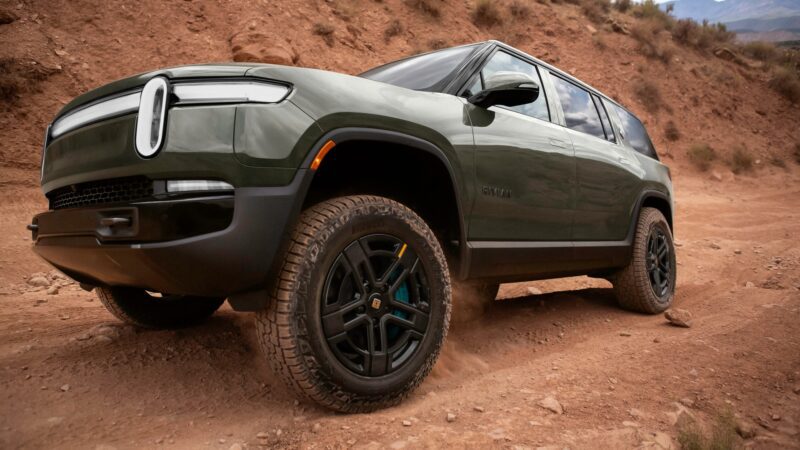 A green Rivian R1S SUV drives on a dirt road, kicking up dust as it navigates a rocky, off-road terrain.