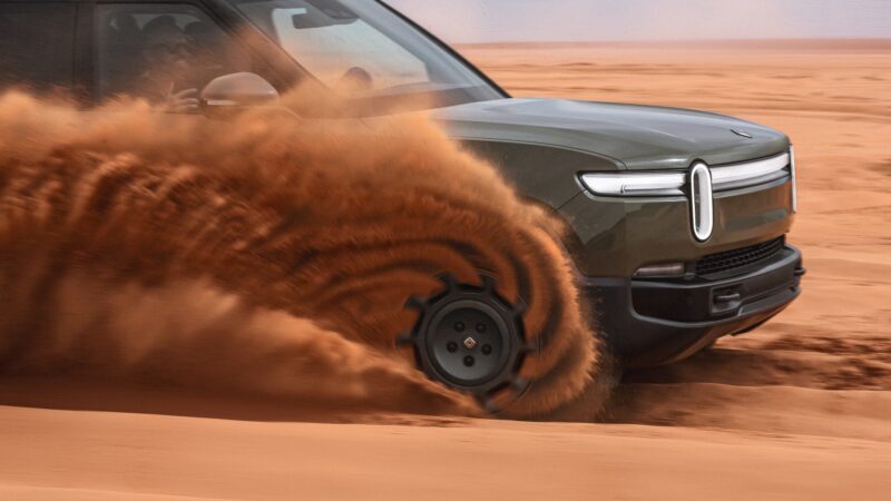 A green Rivian Gen 2 pickup truck drives through orange sand, its front tire kicking up a large spray of dust.