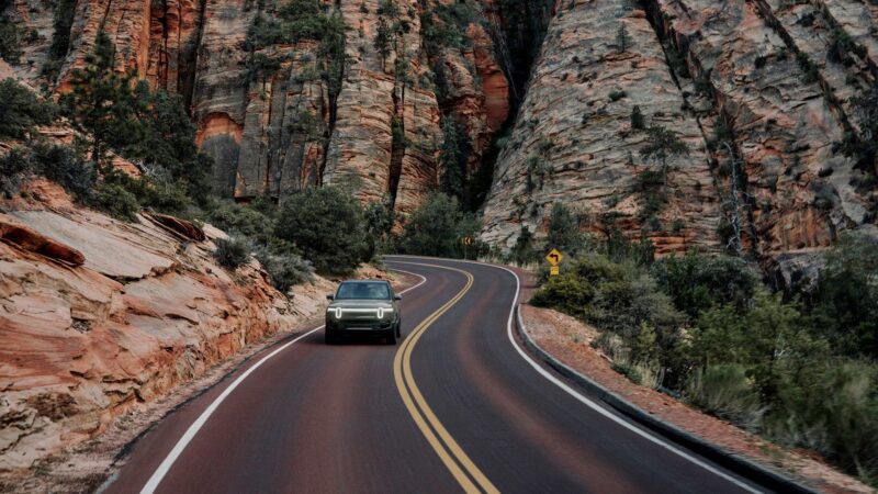 A Rivian Gen 2 R1S drives along a winding road through a rocky canyon with layered cliffs and lush green vegetation on either side.