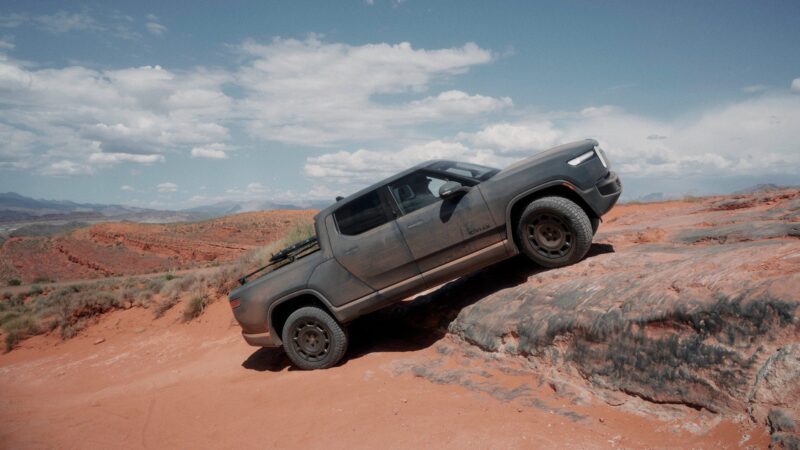 A gray Rivian Gen 2 R1T Quad-Motor pickup truck climbs a rocky, red desert hill under a partly cloudy sky with distant mountains in the background.