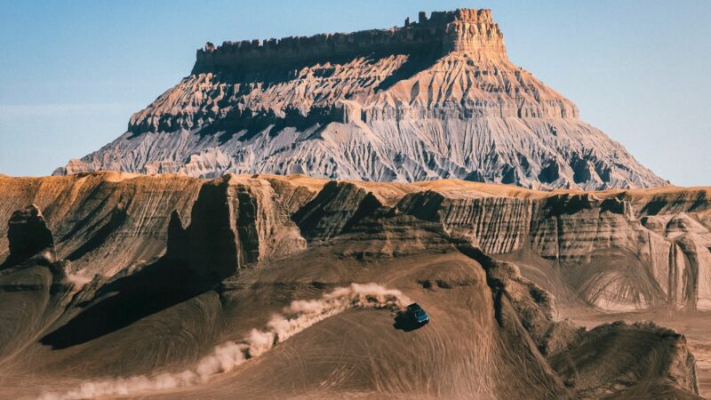 A Rivian Gen 2 R1T drives up a dusty, barren hill in a rugged desert landscape, with a large, flat-topped mesa dominating the background under a clear sky.