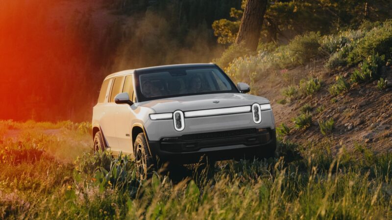 A white Rivian Gen 2 R1S electric SUV drives up a grassy, sunlit hillside with trees and wildflowers in the background.