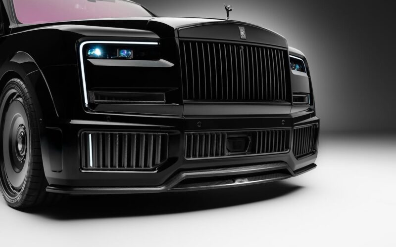 Close-up view of the front of a black Rolls-Royce Cullinan with prominent grille, rectangular headlights, and glossy finish, enhanced by a carbon fiber widebody and striking 24-inch rims in a studio setting.