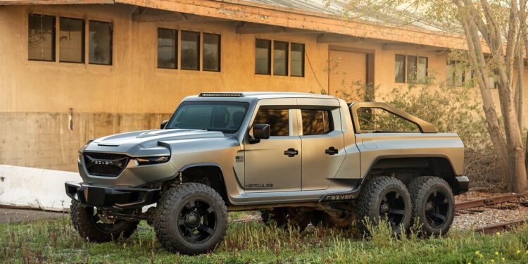 A large, matte grey, six-wheeled pickup truck—a unique creation—is parked outdoors beside a beige industrial building with several windows, hinting at a garage specializing in custom cars for sale.