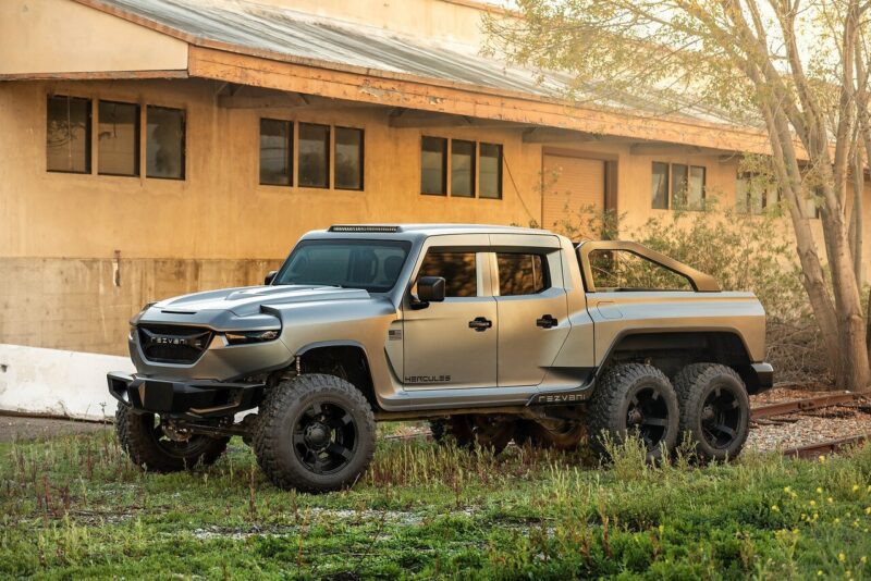 A large, matte grey, six-wheeled pickup truck—a unique creation—is parked outdoors beside a beige industrial building with several windows, hinting at a garage specializing in custom cars for sale.