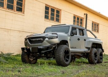 A matte grey six-wheeled Rezvani Hercules pickup truck, one of the most unique custom cars, is parked on grass beside a beige industrial building.