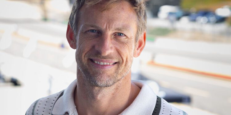 A man with light brown hair and a light beard smiles while standing outdoors on a sunny day, wearing a white and green collared shirt. The blurred road hints at the excitement of the Rolex Monterey Motorsports Reunion with Grand Marshal Jenson Button.