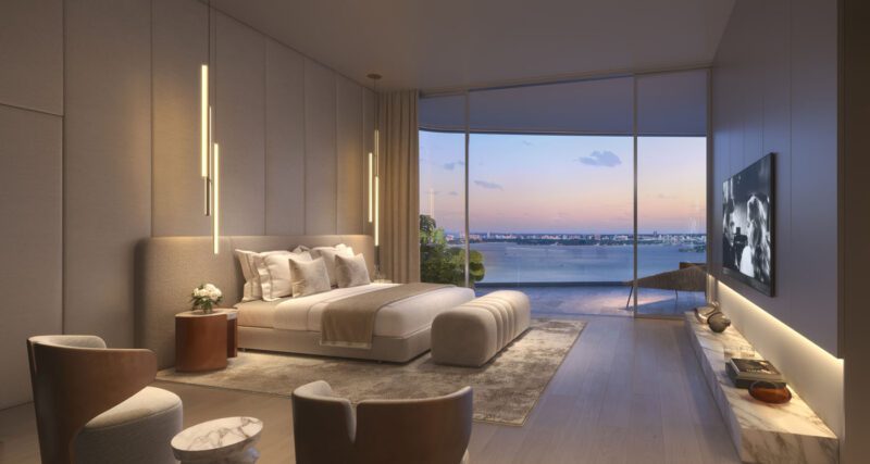Modern bedroom with large bed, two chairs, TV, and floor-to-ceiling windows offering a view of water and city skyline at sunset.