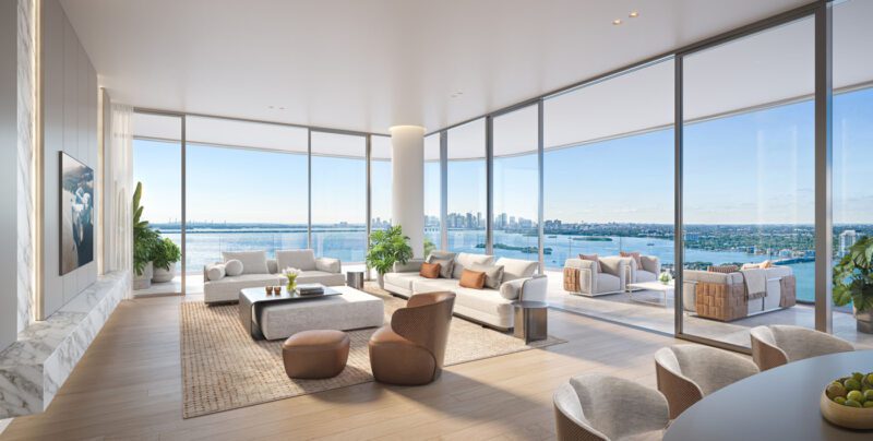 Spacious modern living room with large windows, neutral-toned furniture, indoor plants, and views of the city skyline and water from the balcony.