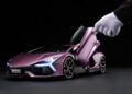 A person wearing a white glove opens the scissor door of a metallic purple Lamborghini Revuelto 1:8 model sports car against a black background.