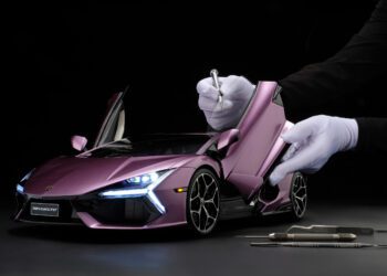 A person in white gloves uses tools to work on a detailed purple Lamborghini Revuelto model with open doors against a black background.