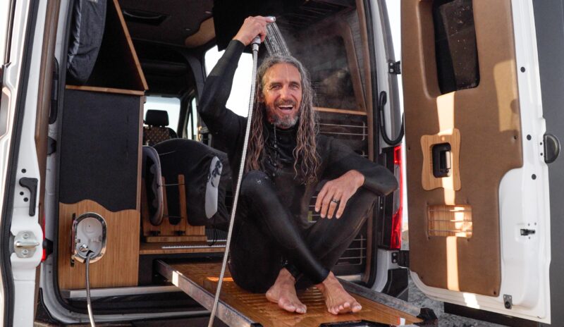 A person in a wetsuit sits on a wooden slide-out platform at the back of a Rogue Van Company van, showering with a hose and smiling, with the van doors open.