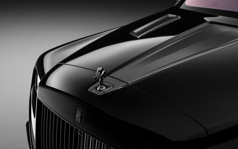 Close-up view of a black Rolls-Royce Cullinan hood featuring the Spirit of Ecstasy ornament, detailed lines, and hints of a carbon fiber widebody under soft lighting.