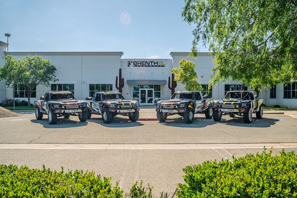 Four off-road race trucks are parked in front of a white building with the sign "Brenthel Industries," ready for an Off-Road Adventure on a sunny day.