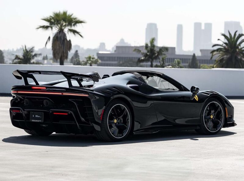 A sleek black SF90 XX Spider with a red carbon fiber rear spoiler is parked on a rooftop, with palm trees and city buildings visible in the hazy background.