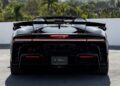 Rear view of a black SF90 XX Spider convertible parked outdoors, featuring taillights, spoiler, and rear grille with Red Carbon Fiber accents. Trees and buildings can be seen in the background near Ferrari Beverly Hills.
