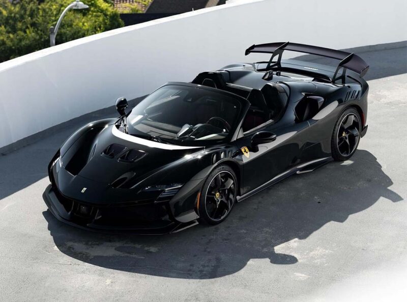 A black Ferrari SF90 XX Spider convertible with an open top is parked on a rooftop with a white curved barrier in the background, highlighting its Red Carbon Fiber accents.