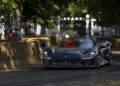 A dark blue Ferrari F80 speeds down the Goodwood track lined with hay bales, as people watch eagerly and a "START" sign looms in the background.