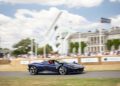 A blue Ferrari speeds past hay bales on the Goodwood racetrack during the F80 Hillclimb, with a large sculpture and historic building in the background under a partly cloudy sky.