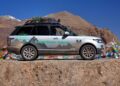 A Range Rover Hybrid prototype parked on a rocky mountain road with snow-capped peaks and colorful prayer flags in the background, echoing the adventurous spirit of Land Rover Classic to showcase 10 historic expedition icons at Pebble Beach 2025.