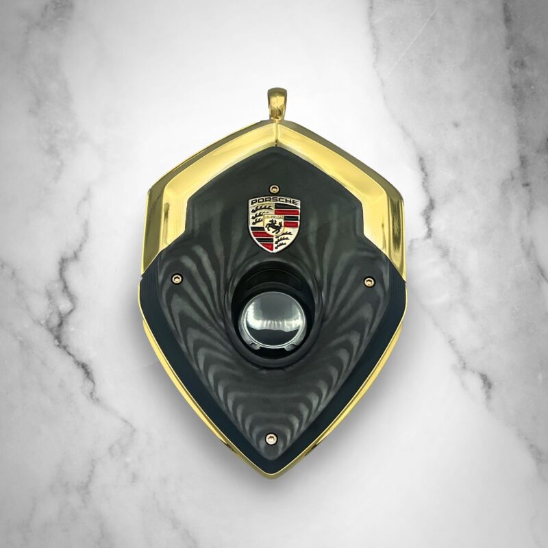 A shield-shaped pendant with gold edges, a black center featuring a lens, and a Porsche logo at the top, displayed on a light marble background—perfect for those looking to upgrade your style with luxury accessories.