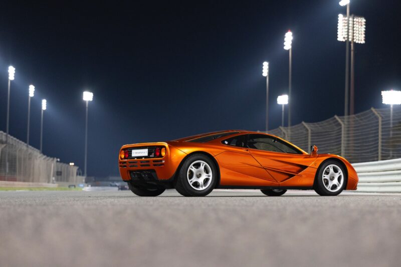 An orange McLaren Road Car Collection sports car is parked on a racetrack at night, illuminated by tall floodlights with fencing visible in the background—a scene reminiscent of Mansour Ojjeh’s passion for performance.