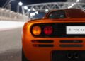 Close-up of the rear end of an orange McLaren Road Car Collection sports car with round tail lights, parked on a racetrack at night with blurred lights in the background.