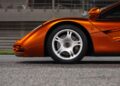 Close-up side view of a McLaren’s front section, highlighting the wheel, tire, and aerodynamic details from the Road Car Collection, with a racetrack barrier in the background.