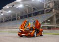 An orange McLaren sports car with its gullwing doors open is parked on a racetrack in front of empty grandstands at night, a striking addition to any Road Car Collection.