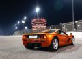 An orange McLaren Road Car Collection sports car is parked on a racetrack at night with bright lights and a tall, illuminated circular building in the background.