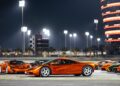 A group of orange sports cars from the McLaren Road Car Collection is parked on a racetrack at night, with bright stadium lights and palm trees in the background.