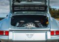 The open rear trunk of a silver classic car reveals a visible engine with quilted interior lining and a distinctive logo on the back panel, echoing the craftsmanship seen in air-cooled 911 restorations by Theon Design.