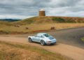 A silver air-cooled 911 sports car is parked on a paved road beside a grassy hill with a small round stone building on top, under a cloudy sky.