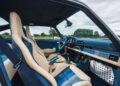 The interior of a sports car featuring beige and blue seats, a blue steering wheel, manual gear shift, and metal foot pedals, with countryside visible through the windows—classic styling inspired by Theon Design’s Air-Cooled 911.