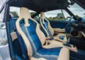Interior of a car showing two sport seats with blue and white plaid fabric and cream leather accents, manual gearshift, and partially open door; trees visible through windows—a bespoke touch reminiscent of a classic Air-Cooled 911 by Theon Design.