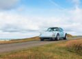 A classic white air-cooled 911 glides along a narrow country road bordered by grass under a partly cloudy sky, embodying the spirit of Theon Design.
