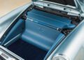 The image shows the front trunk of a silver Theon Design air-cooled 911 with blue upholstery, featuring a geometric logo on the interior panel and a carpeted storage area.