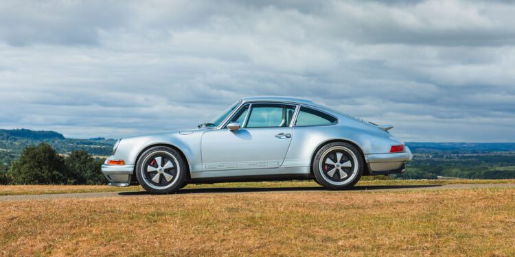 A silver air-cooled 911 coupe by Theon Design is parked on a grassy hill, set against a scenic landscape and cloudy sky.