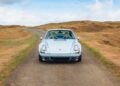 A classic light blue air-cooled 911 Porsche sports car is parked on a narrow paved road surrounded by grassy hills under a partly cloudy sky.