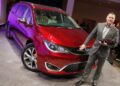 A man in a suit stands next to a red minivan on display indoors, resting one hand on the vehicle's hood and holding an award, embodying the spirit of "Back in the Driver’s Seat: Tim Kuniskis to Lead Stellantis’ Comeback In North America.