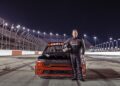 A race car driver in a black racing suit stands holding a helmet in front of a red RAM truck on a lit racetrack at night, echoing the spirit of "Back in the Driver’s Seat: Tim Kuniskis to Lead Stellantis’ Comeback In North America.