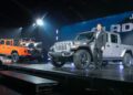 A man in a suit stands next to a gray Jeep Gladiator on stage, as an orange Jeep Gladiator carrying ATVs is displayed in the background—symbolizing Stellantis’ comeback in North America under Tim Kuniskis’ leadership.