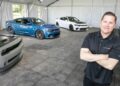 A man stands with arms crossed in front of three Dodge cars—grey, blue, and white—inside a large tent with rows of empty folding chairs, symbolizing Stellantis’ comeback in North America under Tim Kuniskis’ leadership.