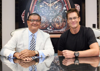Two men sit at a table in front of a large Jacob & Co. Twin Turbo Furious watch image, both wearing wristwatches and glasses; one wears a suit, the other a black t-shirt.