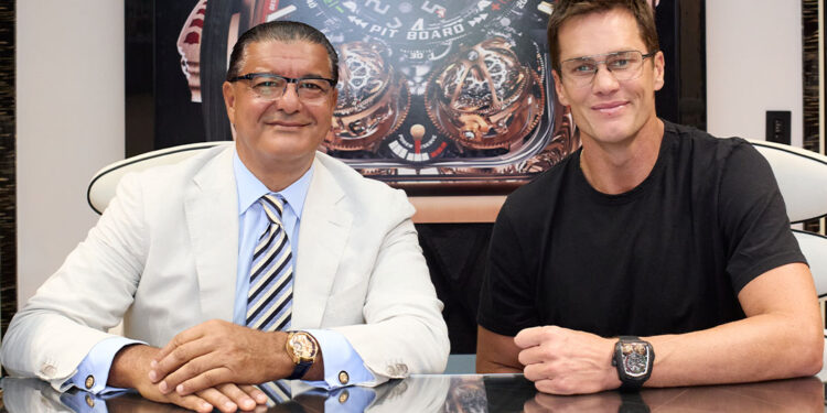 Two men sit at a table in front of a large Jacob & Co. Twin Turbo Furious watch image, both wearing wristwatches and glasses; one wears a suit, the other a black t-shirt.