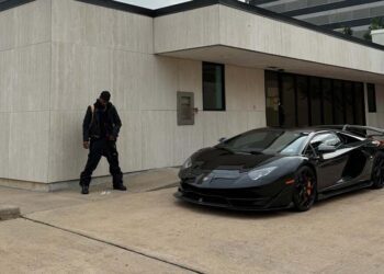 A person dressed in black stands near a beige building, with a sleek black Lamborghini parked nearby on the concrete—channeling the bold style of JACKBOYS 2 and Travis Scott.
