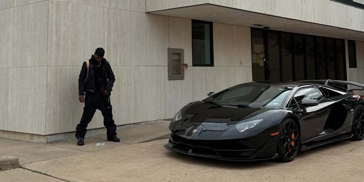 A person dressed in black stands near a beige building, with a sleek black Lamborghini parked nearby on the concrete—channeling the bold style of JACKBOYS 2 and Travis Scott.