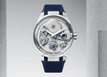 A silver Ulysse Nardin wristwatch with a visible mechanical movement, blue strap, and metallic accents, displayed against a gradient gray background.