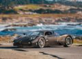 A black Pagani sports car is parked near a coastal road, surrounded by rocky shoreline and greenery under a blue sky, hinting at its next adventure—perhaps hillclimb runs at the Goodwood FoS 2025.